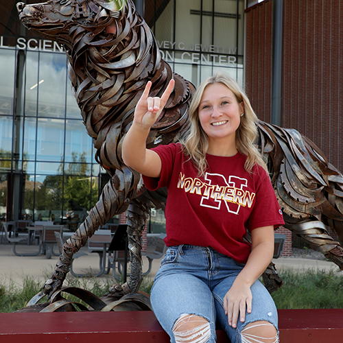 Kinze Stradtman sitting in front of the wolf statue in front of the Jewett Regional Science Education Center and making the wolves hand sign by touching her middle and ring fingers to her thumb and pointing her index and pinkie fingers up