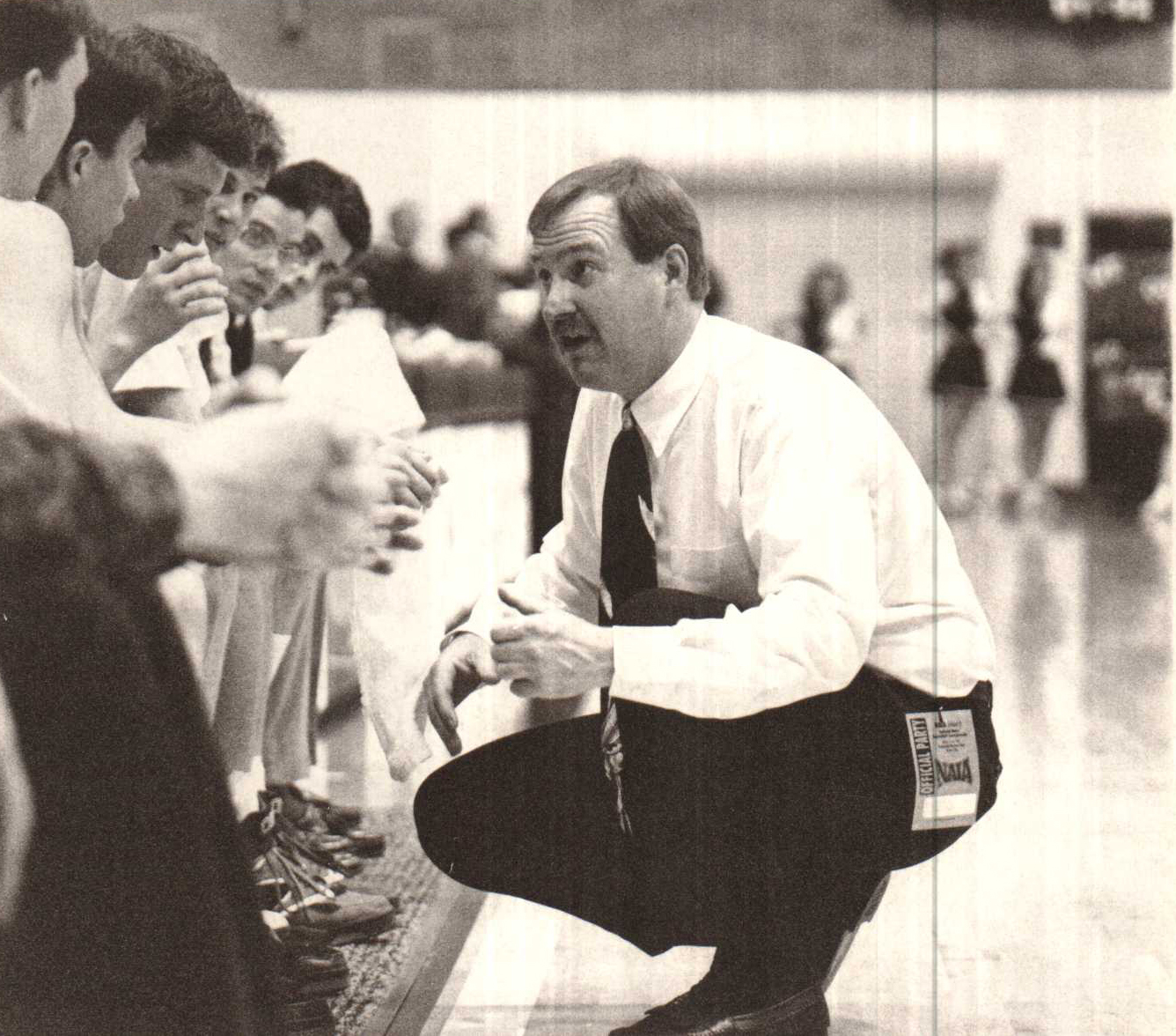 Historical photograph of Bob Olson crouching in a huddle in front of large group of student athletes seated in gymnasium