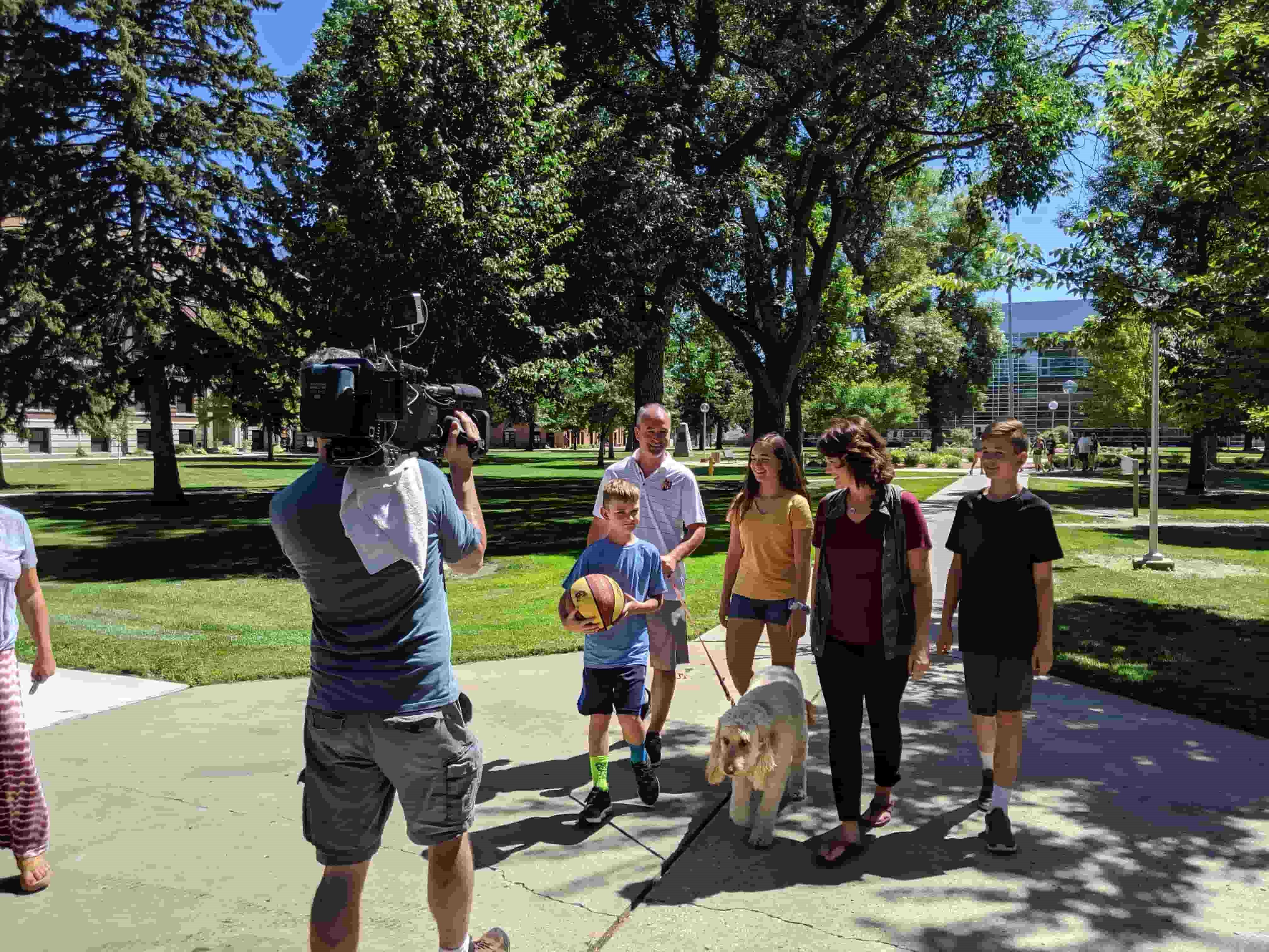 Wolves Men’s Basketball Coach Saul Phillips, his wife, Nicole, their children, and their dog walking along campus green while camera films them for episode