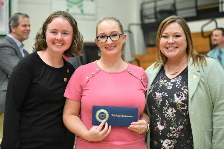Three women smiling at the camera; the middle woman, in a pink shirt and glasses, is holding a certificate for a teaching award with her name, Nichole Bowman, on it
