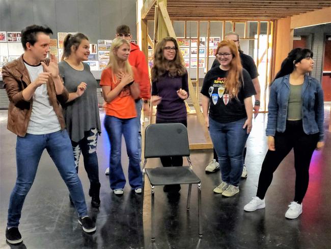 Group of theater students rehearsing together around a chair on stage