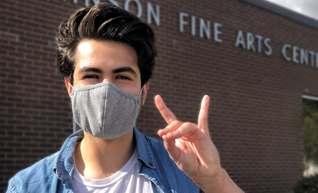 Student wearing mask making NSU Wolf sign in front of campus building