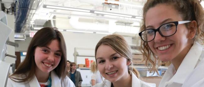 Three female college students in lab coats smiling at camera